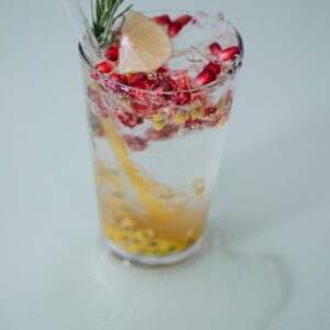 From above glass of tasty fresh cold drink with pomegranate seeds and passion fruit pure decorated rosemary and lemon and added transparent straw on white table in puddle